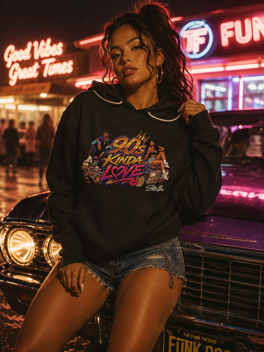 A woman wearing a black graphic hoodie with a colorful retro 90s graphic on the chest, posing in front of a vintage car at night with neon signs in the background.